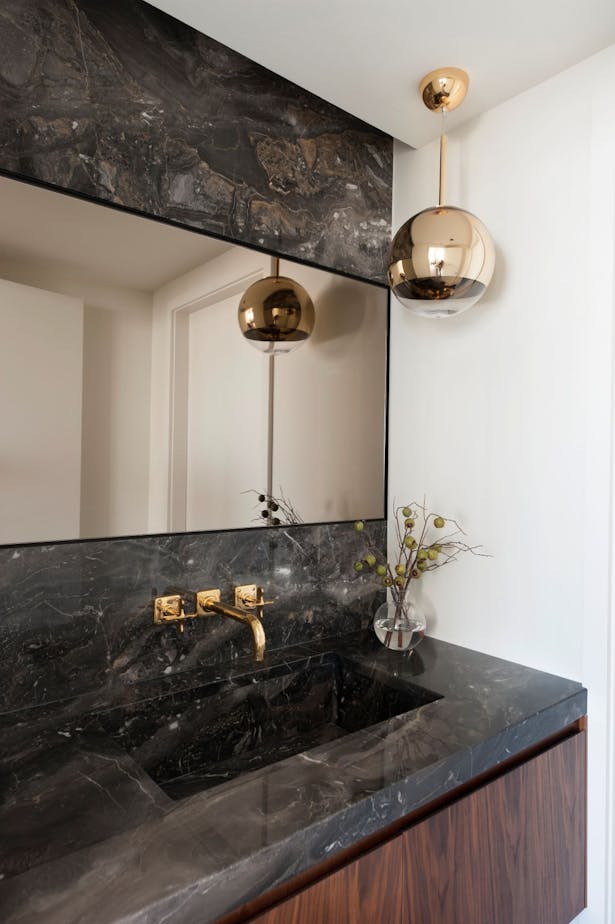 The large powder room is divided into separate zones, with a vanity in the front area. A bronze tinted mirror is set flush with stone panels above and below. Aged brass fittings and lighting were only used at this special guest sink, which was integrated into the stone counter for an upscale experience.